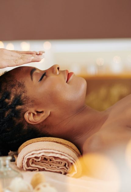 Therapist holding hands over head of pretty young Black woman to transfer energy during healing reiki session