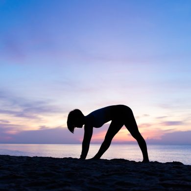 Silhouette of beautiful woman practicing yoga asana on the beach with sunrise on twilight blue vibrant sky and calm sea in background. Piramid pose, Spine, Stretching, June 21, International Yoga Day